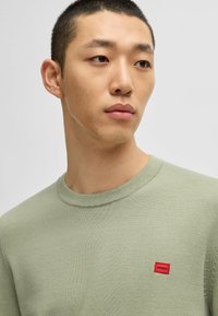 Grüner Strickpullover mit Rundhalsausschnitt und kurzen Ärmeln. Kleines rotes "HUGO"-Logopatch auf der linken Brust, glatte Textur.