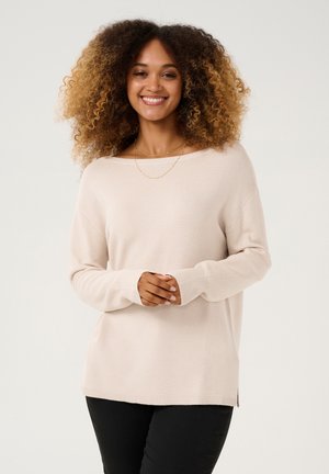 Maglione beige a maniche lunghe con texture a coste, scollatura a barca e spacchi laterali. Abbinato a pantaloni neri.