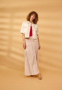 Femme aux cheveux bouclés porte une veste beige à manches courtes, un haut rouge, une longue jupe fleurie et des chaussures tissées beiges sur un fond beige.