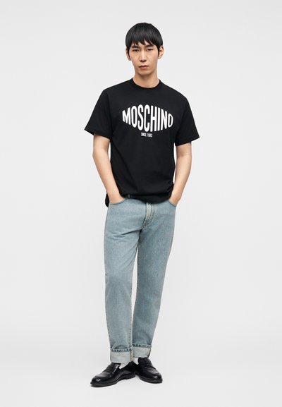 Μαύρο βαμβακερό μπλουζάκι με λευκό λογότυπο "MOSCHINO", σετ με ανοιχτόχρωμο τζιν παντελόνι και μαύρα παπούτσια. Απλή, καθημερινή ενδυμασία.