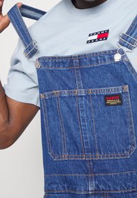 Džíny na kšandy ve středně modré barvě s nastavitelnými proužkovanými popruhy, stříbrnými kovovými doplňky a červenou nášivkou s logem Levi's na kapse. Pod tím je světle modrá košile s krátkými rukávy.