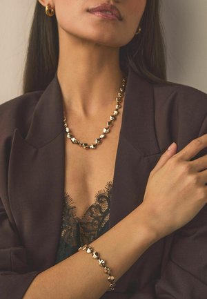 Woman wearing a dark blazer and lace top, accessorized with a gold heart-shaped necklace, bracelet, and hoop earrings.