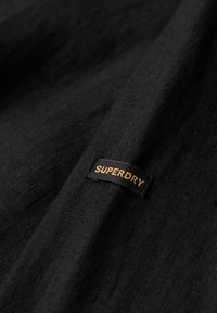 Tela negra con una superficie texturizada; presenta una etiqueta negra con texto dorado que dice "SUPERDRY" en el borde.