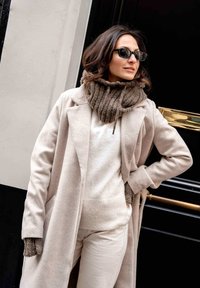Beige wool coat over a cream sweater with ribbing, paired with fingerless knit gloves. Accents include a brown knitted scarf and dark sunglasses.