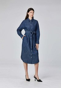 Femme portant une robe en denim à manches longues, boutonnée, avec une ceinture nouée à la taille et des chaussures à talons hauts noires à lanières, se tenant devant un fond uni.