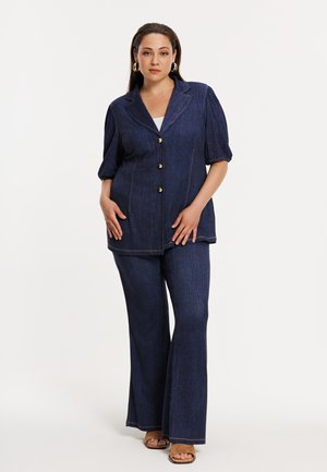 Kvinde iført mørkeblå denimjakke med knapper og pufærmer samt matchende vide bukser, stående med hænderne nær hofterne.