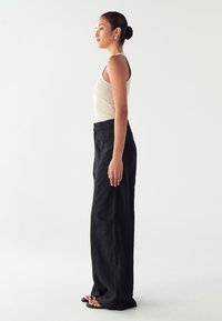 Svetlo bež rebrast majica bez rukava u paru z visoko strukiranim crnim širokim hlačama, glatke teksture i ravnim crnim sandalama, prikazano sa strane.