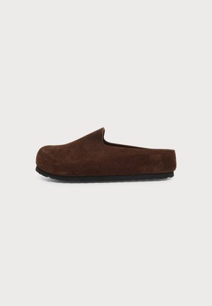Brun ruskind slip-on tøffel med sort sål, vist fra siden på en ensfarvet lysegrå baggrund.