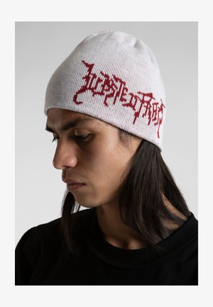 Witte beanie met rode geweven tekst, ribbelstructuur en afgeronde kroon. Licht scheef gedragen door een persoon met lang haar.