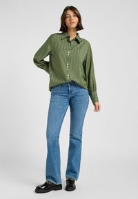 Camicia lunga verde in satin con bottone frontale, abbinata a jeans svasati blu. La camicia presenta una sottile texture a righe.