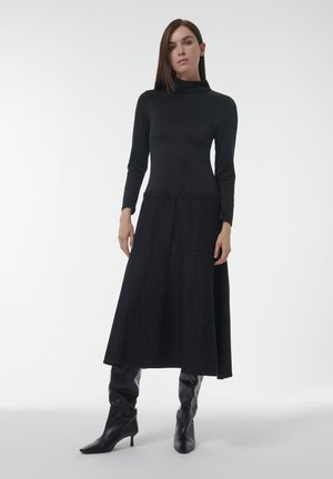 Langes schwarzes Kleid mit eng anliegenden langen Ärmeln und einem hohen Kragen, das in einen weit ausgestellten, dunklen strukturierten Rock übergeht. Tragen mit schwarzen kniehohen Stiefeln.