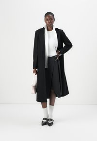 Palton lung negru, bluză albă, fustă gri cu dungi subțiri și curea, șosete albe și pantofi negri cu vârf ascuțit. Geantă de mână roz deschis. Stil minimalist.