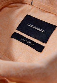 Leinenmischgewebe in Pfirsichfarbe mit schwarzen Etiketten. Oberes Etikett: "LINDBERGH", unteres Etikett: "Leinenmischung", mit weißem Text auf schwarzem Hintergrund.