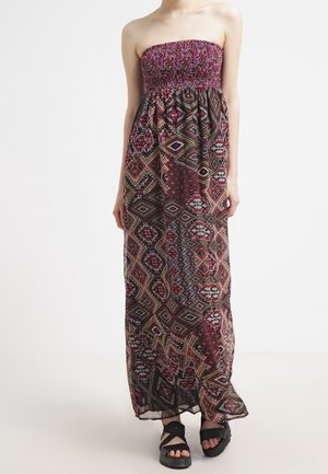 Femme portant une robe longue bustier avec un motif géométrique multicolore et des sandales plateforme noires, debout sur un fond uni.