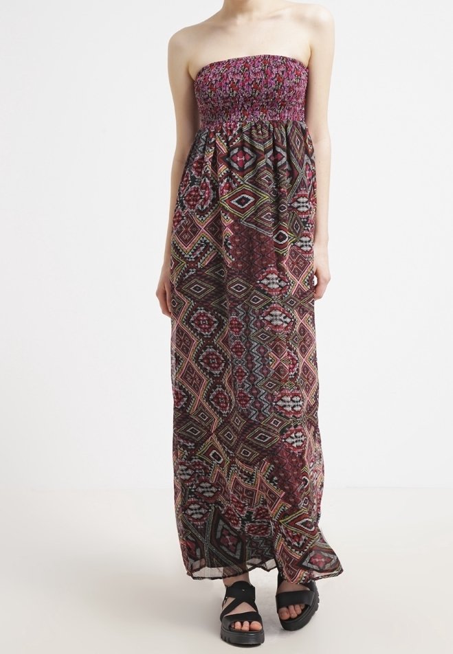 Femme portant une robe longue bustier avec un motif géométrique multicolore et des sandales plateforme noires, debout sur un fond uni.