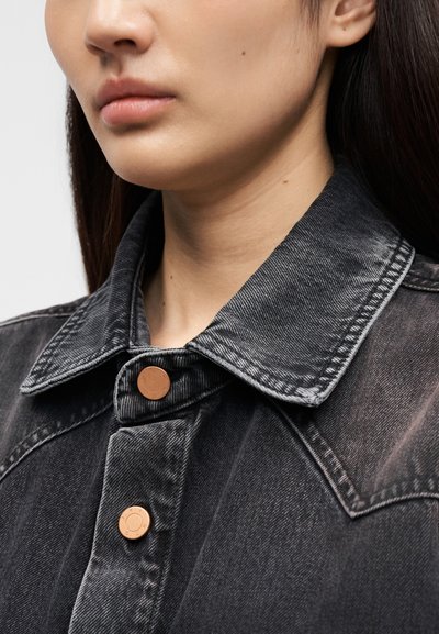 Κοντινό πλάνο ενός ατόμου που φοράει μια ξεθωριασμένη μαύρη τζιν μπουφάν με χάλκινα κουμπιά, που δείχνει το κάτω μέρος του προσώπου, το λαιμό και τις λεπτομέρειες του γιακά.