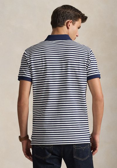 Άνδρας που φοράει μπλε σκούρα και λευκή οριζόντια ριγέ κοντομάνικη πόλο μπλούζα και σκούρο τζιν, βλέποντας από πίσω με ουδέτερο φόντο.