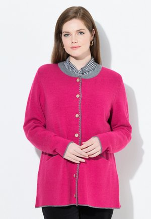 Mujer vestida con un cárdigan de botones rosa brillante con ribete gris sobre una camisa de cuadros en blanco y negro, de pie frente a un fondo blanco.