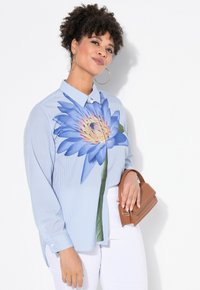 Camicia a righe azzurre con una grande stampa floreale blu sul davanti. Maniche lunghe e colletto con bottoni. Il modello tiene una borsa marrone.