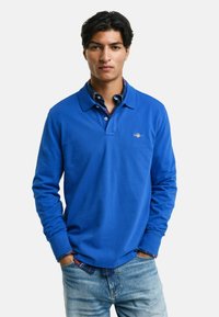 Koninklijk blauwe polo met lange mouwen en een kraag, logo op de borst, en gelaagd met een geruit shirt aan de manchetten, stijlvol gecombineerd met lichte jeans.