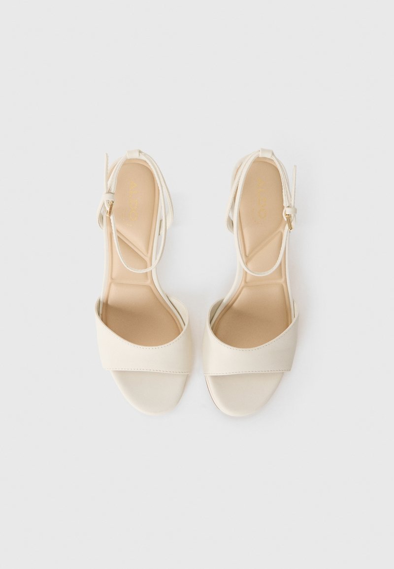 Un par de sandalias de tiras al tobillo de color crema, con punta abierta y tacones bajos, exhibidas sobre un fondo blanco liso.