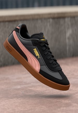 Czarny sneaker Puma z różowym paskiem, brązową podeszwą, złotym logo Puma i czarnymi sznurowadłami unoszący się nad kamienną powierzchnią.