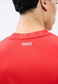 Άνδρας με κοντά μαύρα μαλλιά που φοράει κόκκινο ριμπ πουκάμισο με την επιγραφή "TÜRKİYE" σε λευκά γράμματα στο πάνω μέρος της πλάτης.