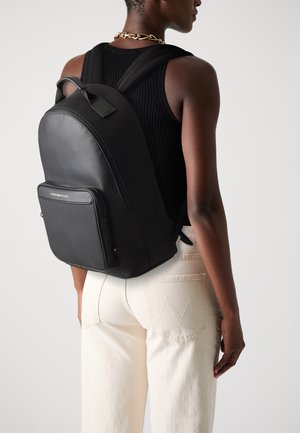 Black leather backpack featuring a front zip pocket, adjustable straps, and a sleek, structured design. Worn on a light-colored outfit.