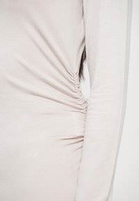 Gros plan sur un vêtement beige à manches longues avec un détail froncé à la taille porté par une personne contre un fond uni.