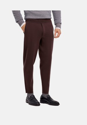 Man draagt donkerbruine op maat gemaakte broek, grijze sokken, zwarte loafers, grijze trui en wit overhemd, handen in zakken op witte achtergrond.