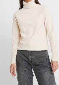 Kremowy sweter z golfem z prążkowanym dekoltem i szwami na ramionach, z długimi rękawami i teksturowanymi akcentami. Połączony z ciemnoszarymi spodniami.