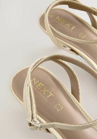 Beige sandaler med korslagda remmar i texturerat material. Guldpräglad logotyp på innersulan. Mjuk, vadderad sula för komfort.