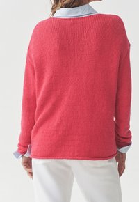 Gestrickter Pullover in leuchtendem Korallenrot, mit lockerem Schnitt und langen Ärmeln, über einem hellblauen Hemd mit Kragen getragen, vor einem neutralen Hintergrund.
