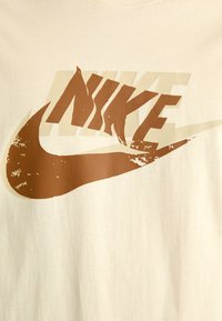Kreminės spalvos medvilninis marškinėliai su dideliu rudos spalvos Nike logotipu ir šonine tekstūros efektu, pateiktu prieš tekstūruotą foną.