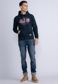 Marine hoodie met een voorzak en een rode grafische print, gecombineerd met blauwe denim jeans en tan kleurige veterschoenen. Zacht materiaal en een ontspannen pasvorm.