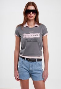 Pilkas trumpas marškinėliai su ro�žiniu apykakle, ant kurių yra juodas "Big Star" logotipas. Sukomponuoti su šviesiai mėlynomis džinsinėmis šortais ir juodu diržu.