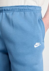 Tuvumā redzamas zilas Nike sporta bikses ar elastīgu jostasvietu un roku sānu kabatā uz baltas fona.