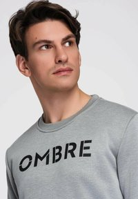 Młody mężczyzna o ciemnych włosach, ubrany w szarą bluzę z nadrukiem "OMBRE" w pogrubionych czarnych literach, spoglądający w zamyśleniu w bok.