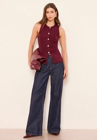 Top bordeaux senza maniche con trama a coste e bottoni dorati, abbinato a jeans denim scuro a gamba larga e a una clutch bordeaux lucida.