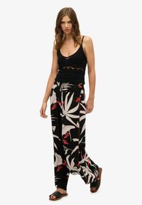 Top de punto negro sin mangas con escote en V, combinado con pantalones de pierna ancha que presentan un estampado de hojas tropicales en blanco y rojo. La modelo lleva sandalias negras.