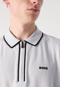 Polo bianco con dettagli neri, dotato di colletto a zip e logo "BOSS" in nero. Tessuto morbido e design strutturato.