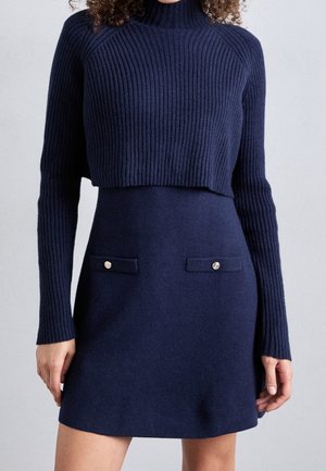 Femme portant un pull court en maille côtelée bleu marine et une jupe taille haute assortie avec des poches boutonnées à l'avant sur un fond uni.