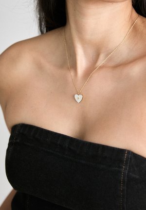 Kvinde iført en guldkæde med en hvid hjerteformet vedhæng, klædt i sort stropfri top, mod en neutral baggrund.