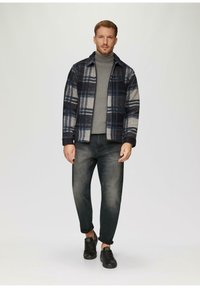 Geruite jas in marineblauw, crème en blauwe kleuren, gelaagd over een grijze coltrui, gecombineerd met een donkere denim jeans en zwarte sneakers.