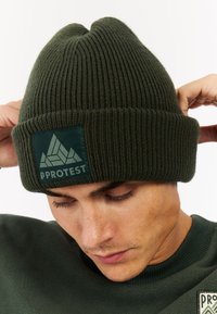 Zelená pletená čepice s žebrovanou strukturou a přehnutým lemem, která má zelenou látkovou nášivku s geometrickým logem a názvem značky.