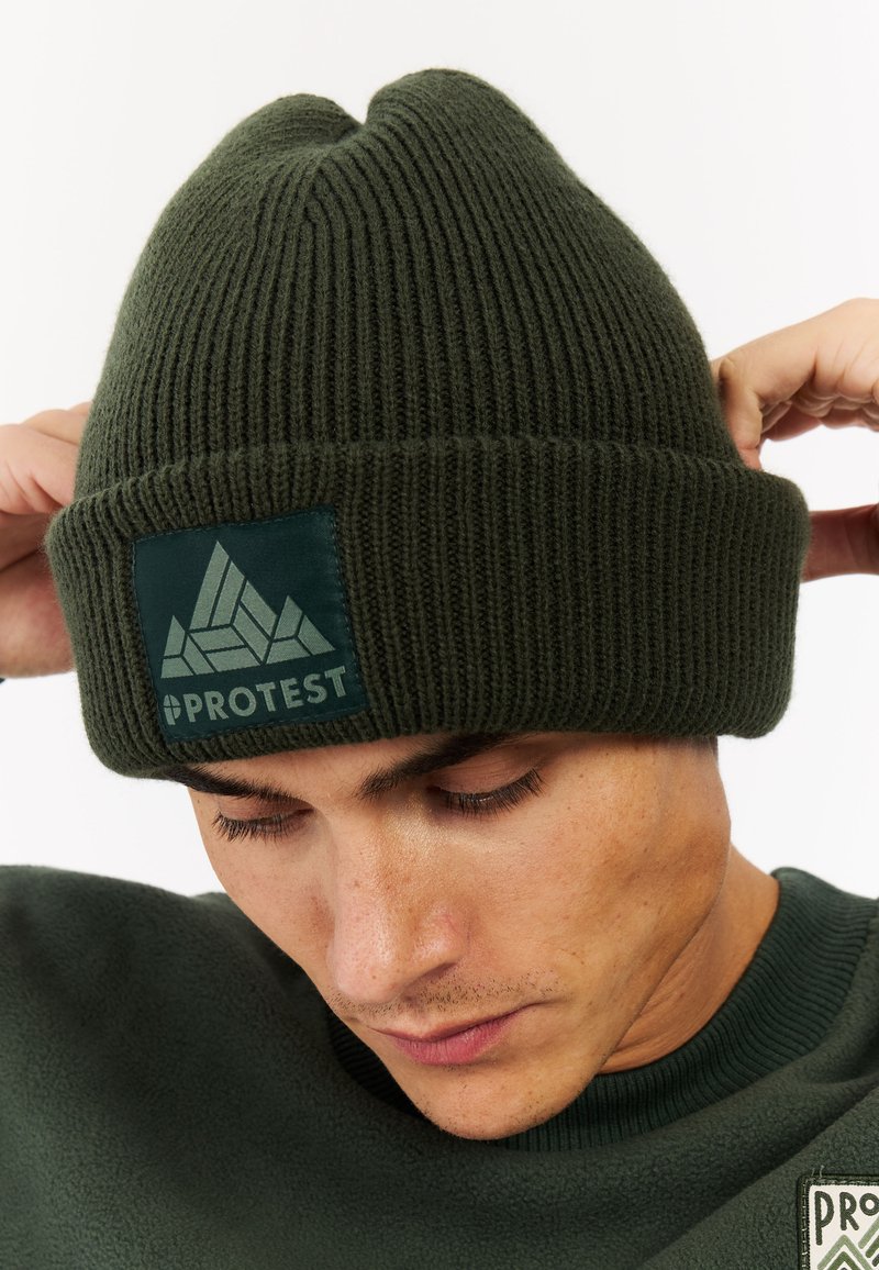 Zelená pletená čepice s žebrovanou strukturou a přehnutým lemem, která má zelenou látkovou nášivku s geometrickým logem a názvem značky.