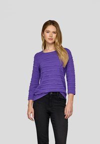 Lila langärmliges Oberteil mit einem strukturierten, geschichteten Design, kombiniert mit schwarzen Jeans. Der Stoff wirkt weich und leicht dehnbar.