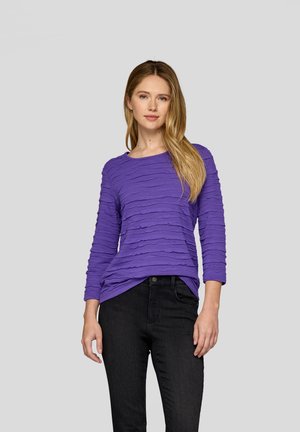 Lila langärmliges Oberteil mit einem strukturierten, geschichteten Design, kombiniert mit schwarzen Jeans. Der Stoff wirkt weich und leicht dehnbar.