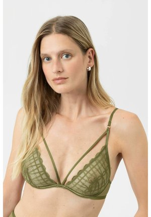 Frau mit langen blonden Haaren, die einen olivgrünen Spitzen-Bralette mit dünnen Trägern trägt, vor einem einfarbigen hellgrauen Hintergrund.