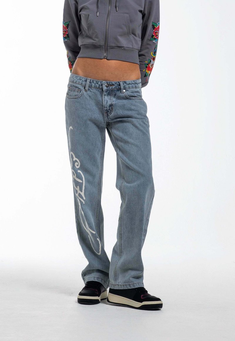 Jeans de mezclilla azul claro con un diseño gráfico blanco en la pierna izquierda, corte recto, combinados con un sudadera corta gris y zapatillas negras.